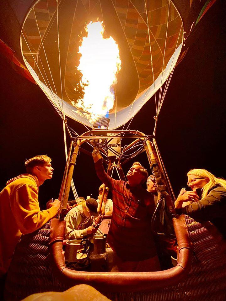A pilot firing the burner inside a glowing balloon basket at night while passengers look up