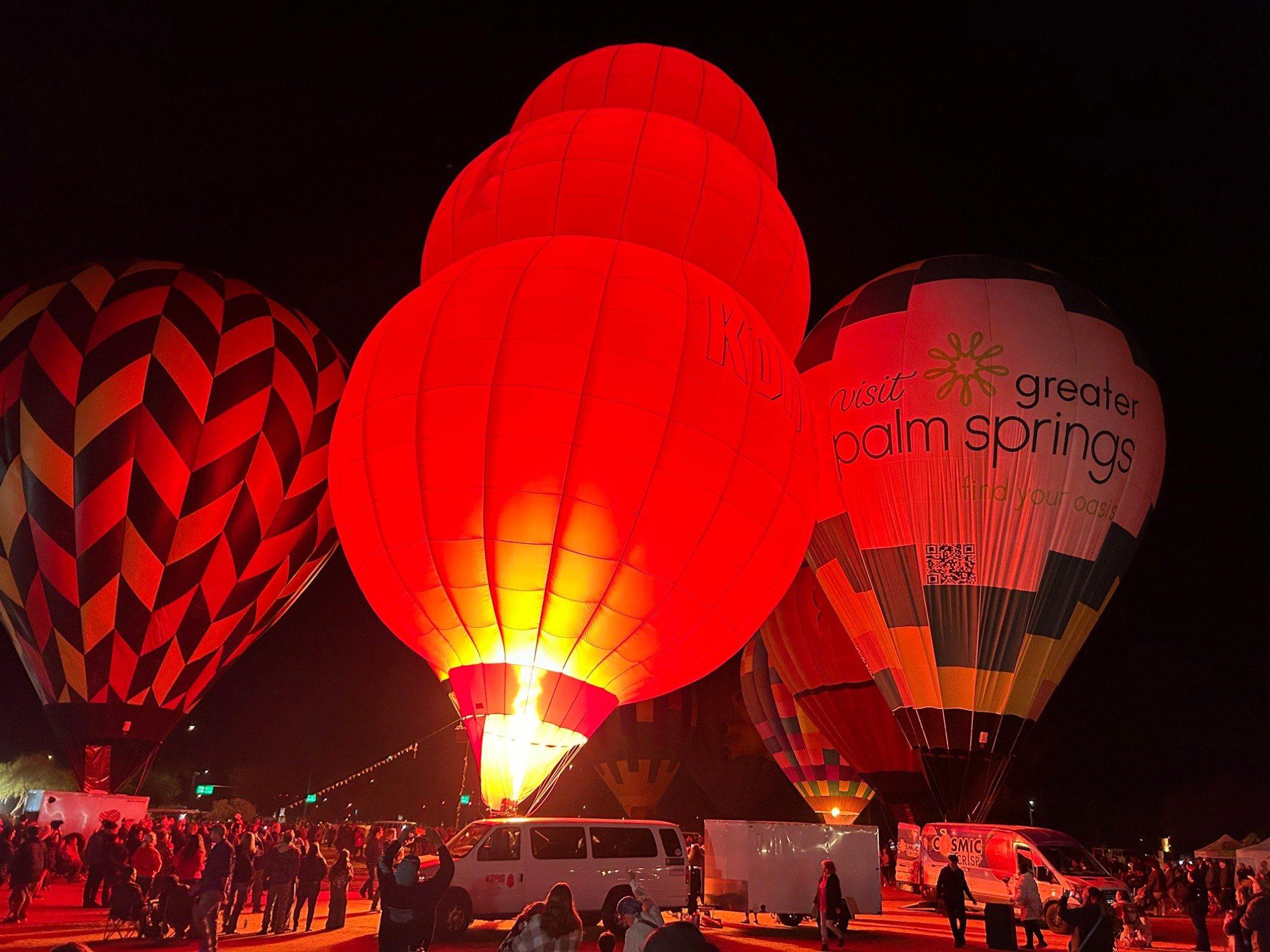 A tall red hot air balloon glows bright as its burner fires during an Evening Desert Glow, with crowds gathered among glowing balloons