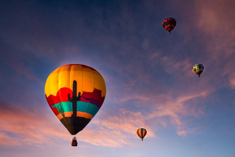 A balloon with a saguaro and desert sunset design rising into a pink morning sky with other balloons in the distance