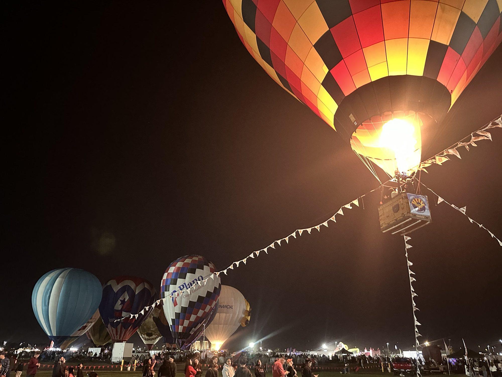A tethered hot air balloon lit by its burner at night, held by flagged tether lines above the crowd, with more balloons glowing on the field
