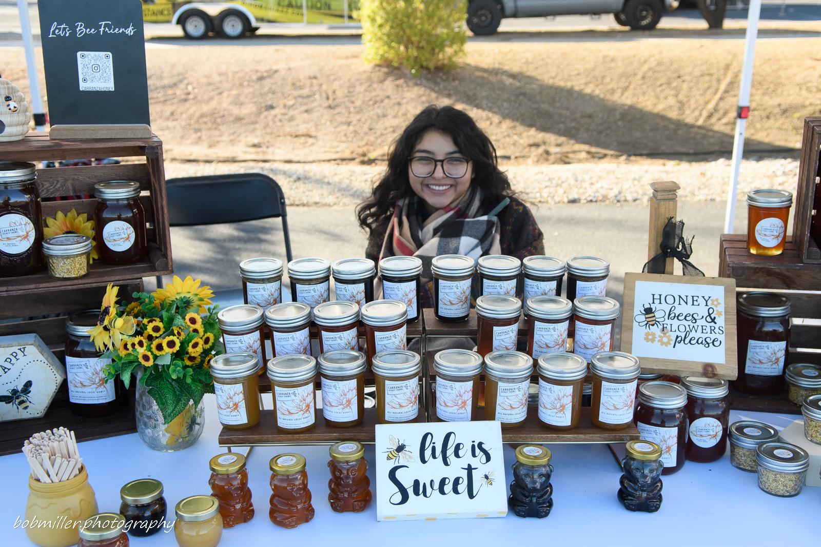 A local vendor smiling behind a table of honey jars and sunflowers at their festival booth