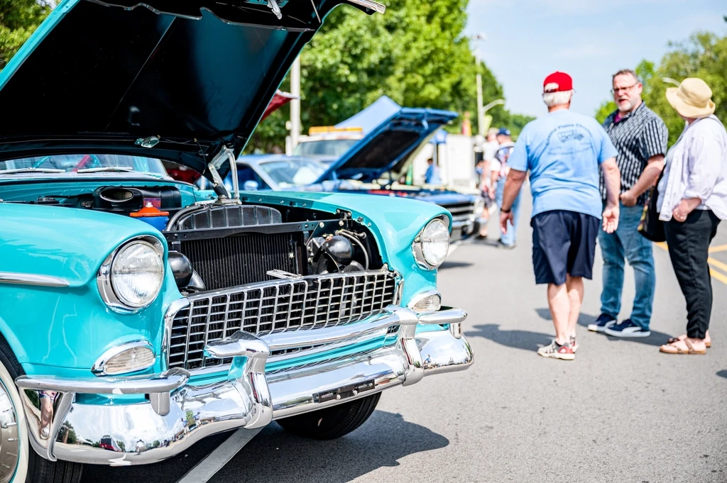 Guests walking past a row of polished classic cars with their hoods open at an outdoor car show