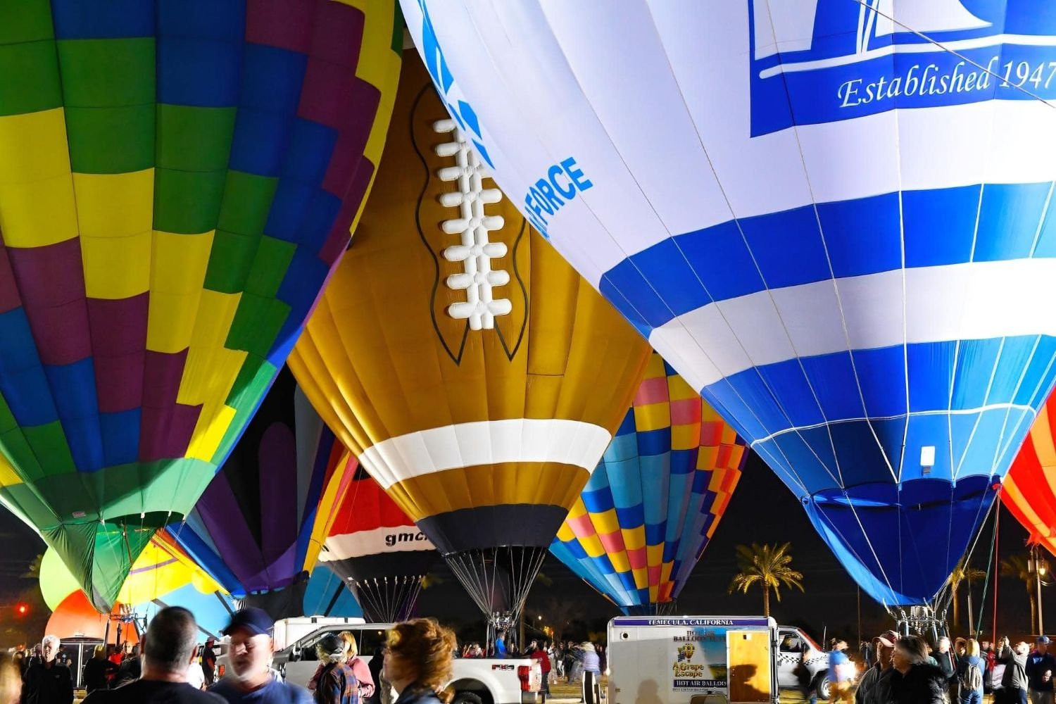 Balloons glowing side by side during an Evening Desert Glow, including a football-shaped balloon, as a crowd gathers on the field