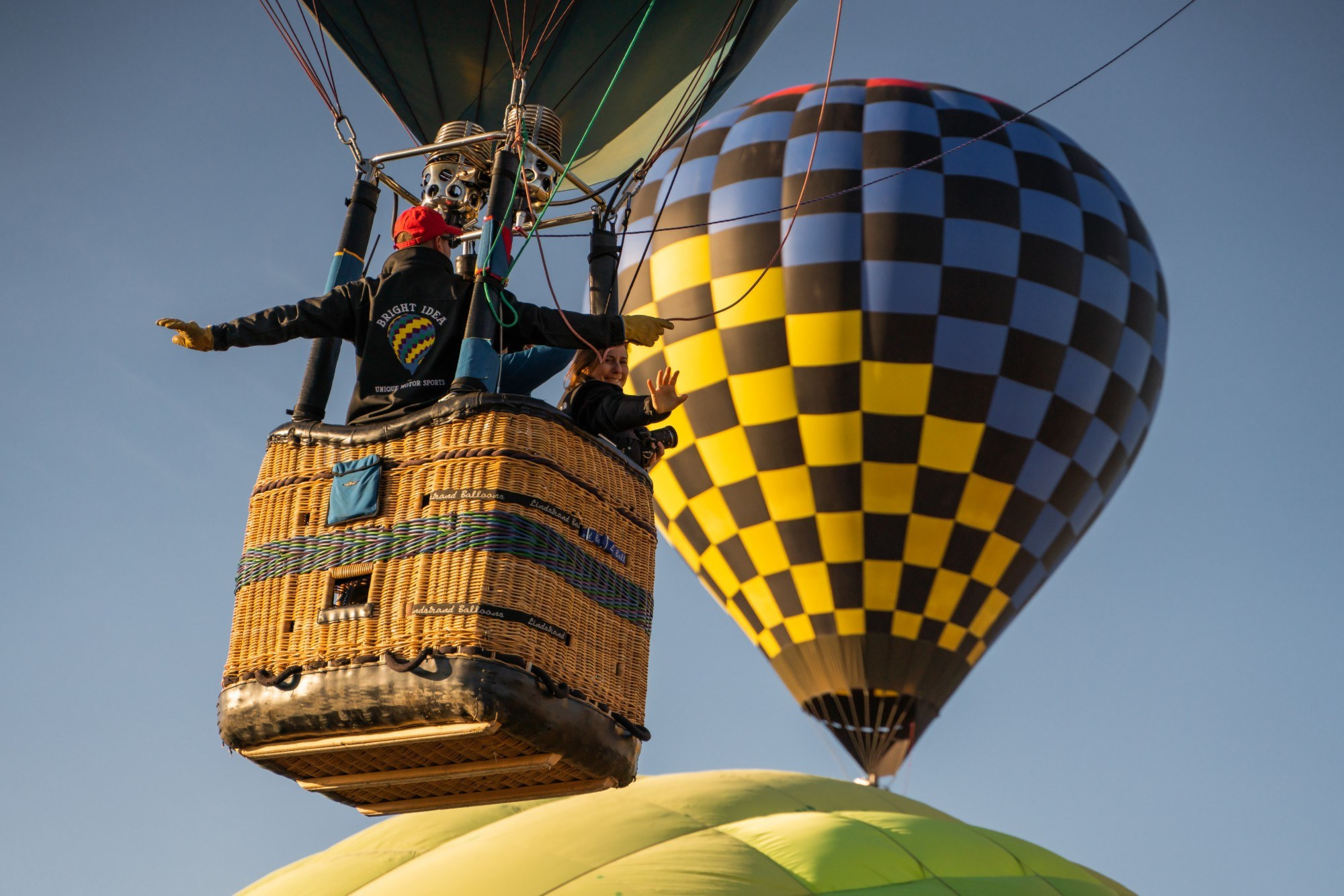 A pilot and a smiling passenger wave from a wicker balloon basket as it lifts off, with a checkered balloon behind them
